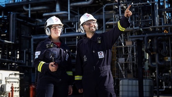 Un hombre y una mujer con cascos blancos, gafas de seguridad y uniformes azules en una planta industrial. El hombre señala hacia arriba y la mujer sonríe