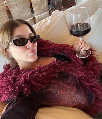 Mujer recostada en un sofá blanco, viste gafas de sol oscuras, abrigo rojo peludo, top rojo oscuro y sostiene una copa de vino tinto