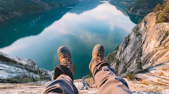 Primer plano de pies con botas de montaña colgando sobre un acantilado rocoso. Abajo, un lago de aguas azules rodeado de montañas boscosas.