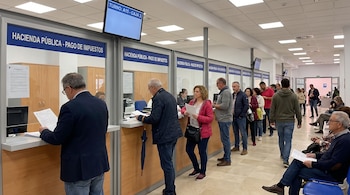 Interior de oficina con personas haciendo fila y en ventanillas. Carteles azules dicen Hacienda Pública - Pago de Impuestos. Un monitor muestra 'TURNO: A15 - CAJA 3'.