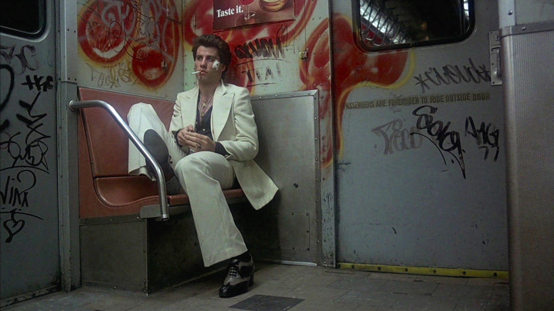 El actor John Travolta interpreta a Tony Manero, fumando un cigarrillo en un vagón de metro lleno de grafitis en una escena clásica de la película Fiebre de sábado por la noche. (Captura de video)