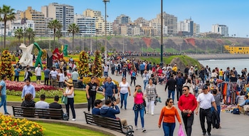 Vista aérea de un concurrido parque en Miraflores, Lima, con multitud de personas paseando entre jardines floridos, esculturas, palmeras, el océano y edificios de la ciudad.