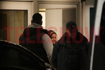Tres personas: una mujer con cabello rubio en el centro sonríe, un hombre de espaldas a su izquierda, un hombre con gorra negra a su derecha. Fondo difuso y oscuro