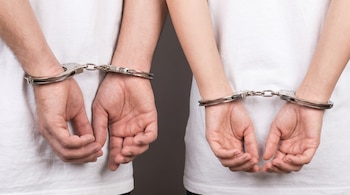 Primer plano de los torsos de un hombre y una mujer, ambos con camisetas blancas, esposados por las muñecas con las manos detrás de la espalda, sobre un fondo gris liso.