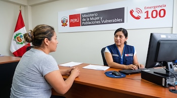 Dos mujeres sentadas a un escritorio con una computadora. Detrás, la bandera de Perú y el letrero del Ministerio de la Mujer y Poblaciones Vulnerables.