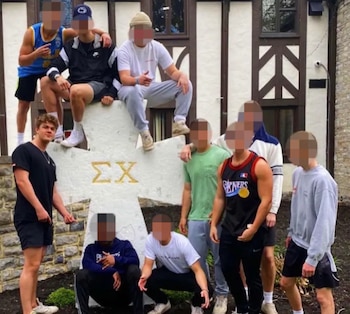 Un grupo de jóvenes rodea una gran cruz de piedra con las letras griegas Sigma Chi frente a un edificio