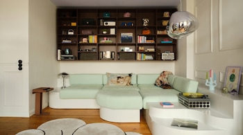 Sala de estar moderna con sofá seccional verde menta, estantería de madera oscura, mesa de centro blanca y lámpara colgante metálica. El suelo es de madera.