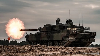 Tanque militar K2 Black Panther camuflado dispara su cañón con gran fogonazo, sobre terreno de tierra. Hay árboles, cielo nublado y un marcador rojo