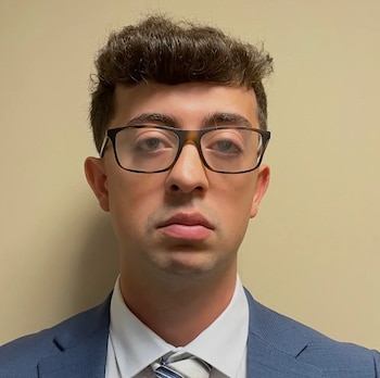Un hombre joven con anteojos de marco oscuro, traje azul, camisa blanca y corbata frente a una pared de color beige