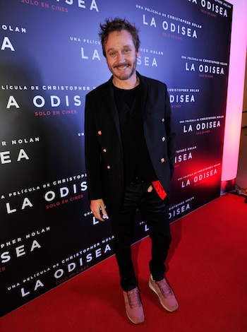 Un hombre sonriendo con barba, vestido con traje negro y zapatillas rosas, de pie en una alfombra roja frente a un telón con texto