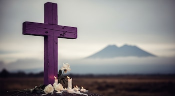 Cruz de madera pintada de color morado con varias rosas blancas y velas encendidas en su base. Al fondo, una silueta montañosa bajo un cielo nublado.