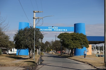 Vista frontal del arco de bienvenida azul que dice 'CIUDAD AMPLIACION FERREYRA'. Una calle asfaltada, árboles y un hombre caminando a la derecha