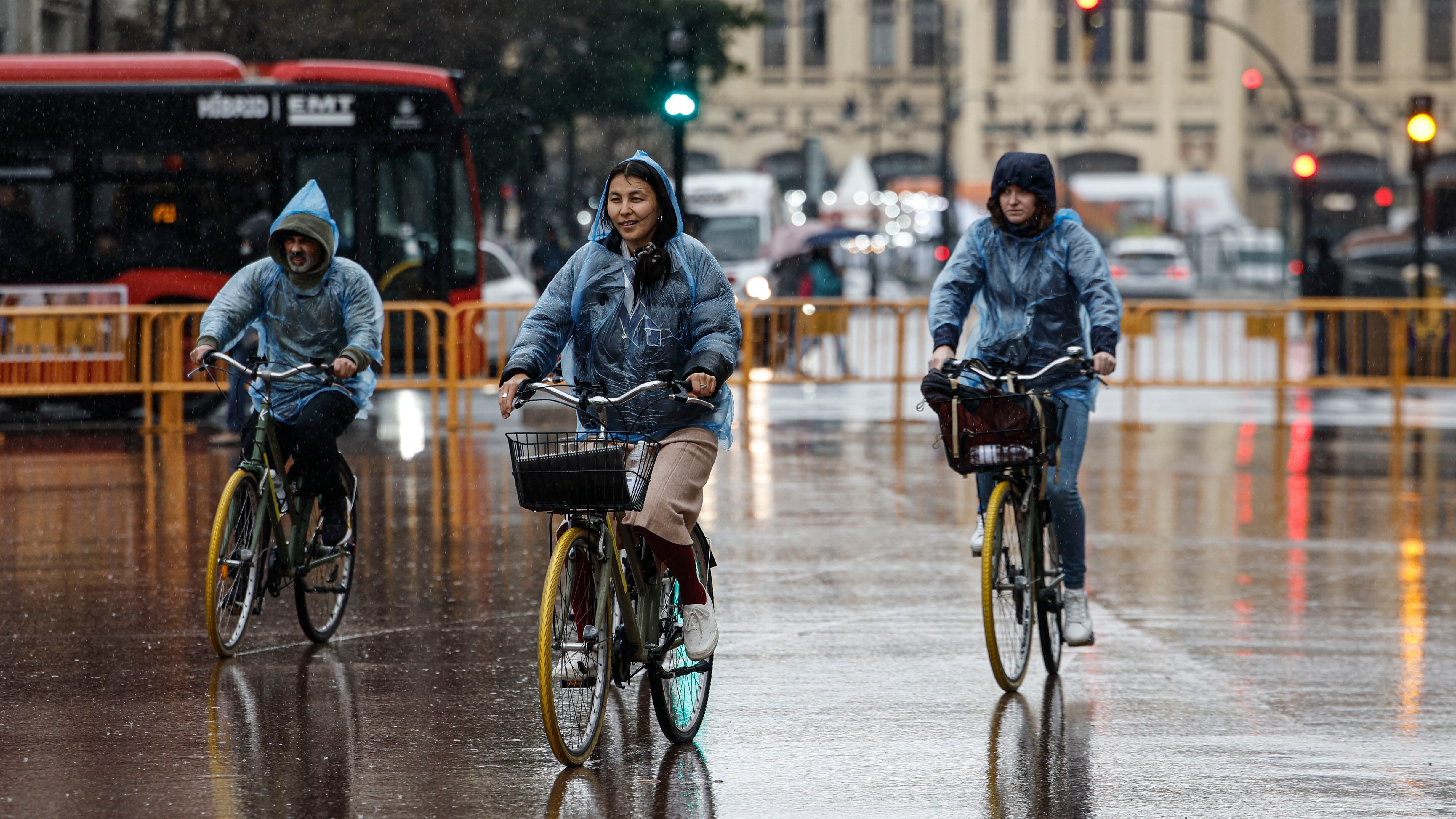 Varias personas en bici se protegen de la lluvia con chubasqueros en Valencia. (Biel Aliño/EFE)
