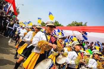 Banda escolar con uniformes blancos, amarillos y negros, sombreros con plumas, tambores y pompones dorados desfila en una calle con público