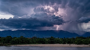 Un rayo ilumina nubes oscuras de tormenta sobre una cadena de montañas con árboles y agua en primer plano.