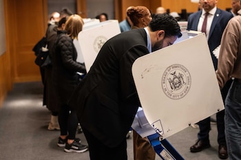 Un hombre de traje negro y camisa azul vota en una cabina electoral blanca con el escudo de Nueva York en una sala con otras personas