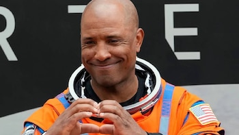 Primer plano de Victor Glover, un hombre calvo con traje espacial naranja y azul, sonriendo y haciendo un corazón con las manos