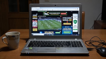 Un ordenador portátil sobre una mesa de madera con su pantalla encendida mostrando un partido de fútbol, una página web con anuncios, una taza y un ratón.