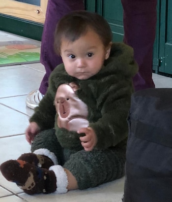 Un bebé con un mameluco verde afelpado con la cara de un mono y calcetines marrones con detalles blancos se sienta en un piso de baldosas