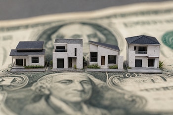 Cuatro pequeñas maquetas de casas con grietas en las paredes blancas, colocadas sobre un billete de un dólar de Estados Unidos.