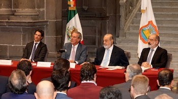 Cuatro hombres con traje y corbata sentados tras una mesa roja con la bandera de México al fondo y varias personas en primer plano.