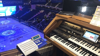 Primer plano de un órgano digital de madera con dos teclados, controles y pantalla, junto a un equipo electrónico azul, sobre la pista de hockey iluminada en un estadio