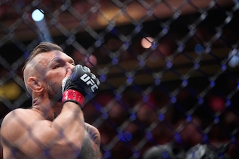 Un hombre con barba, corte de cabello rapado y tatuajes, cubriendo parte de su rostro con un guante negro de UFC, observado a través de una malla metálica de alambre
