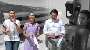 Tres personas, dos mujeres y un hombre, cortan una cinta blanca con tijeras y un bolígrafo; al fondo, un edificio, montañas y un robot.