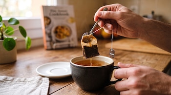 Una mano sostiene una cuchara y el hilo de una bolsa de té negro sobre una taza de cerámica en una mesa de madera.