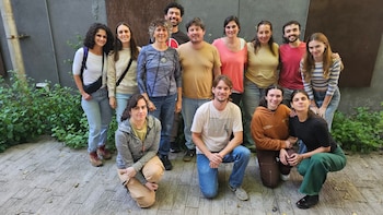 Un grupo de catorce personas, once de pie y tres agachadas en primer plano, sonríen a la cámara en un espacio exterior con una pared oscura y plantas verdes de fondo