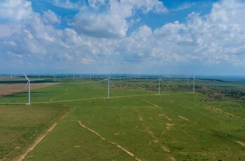 Vista aérea de un vasto campo de molinos de viento blancos en una pradera verde bajo un cielo azul parcialmente nublado, con más turbinas en el horizonte