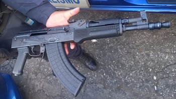 Un oficial de policía sostiene un fusil de asalto negro tipo AK con un cargador extendido, contra un fondo de asfalto y un vehículo azul