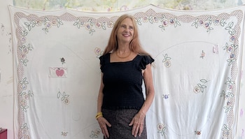 Mujer de cabello largo, blusa negra y falda oscura sonríe ante un textil blanco bordado con motivos florales y un aplique de manzana rosa
