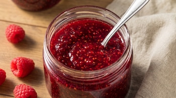 Frasco de vidrio con mermelada roja y una cuchara en su interior, junto a varias frambuesas frescas sobre una mesa de madera.