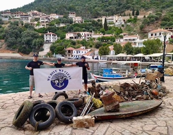 Tres personas sostienen una pancarta blanca frente al mar, rodeadas por varios neumáticos usados y chatarra en un muelle con barcos