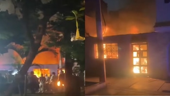 Vista nocturna de una casa en llamas con fuego intenso saliendo de las ventanas y el techo, mientras personas observan desde la calle y un árbol oscuro se alza en primer plano