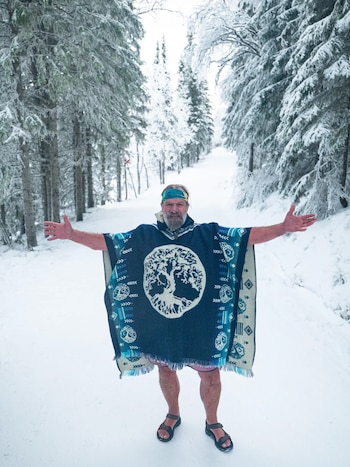 Un hombre barbudo, Wim Hof, con un poncho azul, shorts rosas y sandalias, de pie en un sendero cubierto de nieve con los brazos extendidos, entre árboles nevados