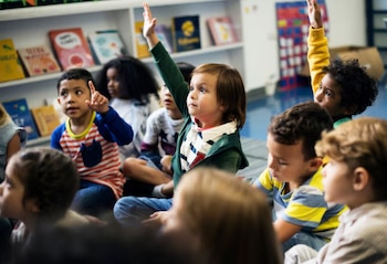 Un grupo de niños de diversas etnias sentados en el suelo de un aula de kindergarten. Tres niños levantan sus manos para participar