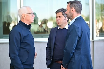 Tres hombres conversando: uno con chaqueta azul y gafas a la izquierda, uno con blazer y suéter azul en el centro, y uno con barba y traje azul a la derecha