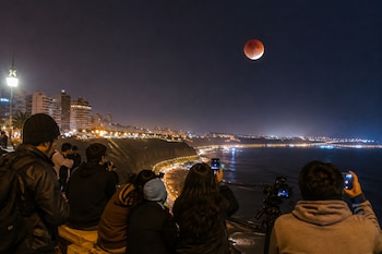 Varias personas de espalda fotografían una luna de tono rojizo en el cielo nocturno sobre la costa iluminada