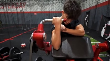 Un joven con camiseta negra realiza curls de bíceps en una máquina roja en un gimnasio; se ve una barra con pesas en el suelo y una red al fondo