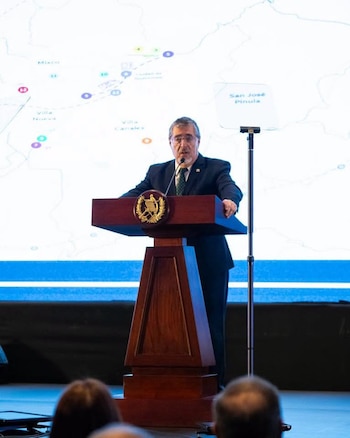 Hombre con traje y corbata habla en un podio de madera con micrófono frente a una pantalla con un mapa de ubicaciones y puntos de colores, una audiencia en primer plano