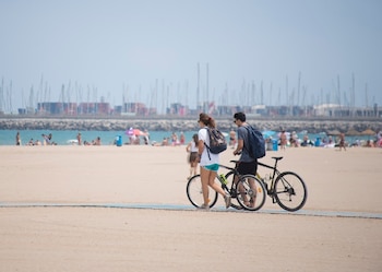 Una pareja joven camina junto a sus bicicletas por la playa de la Malvarrosa, en Valencia, con varios bañistas al fondo.
