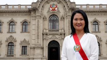 Una mujer con blusa blanca y banda presidencial roja y blanca con un escudo. Detrás hay un edificio de piedra de estilo clásico con ventanas y balcones.