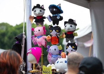 Puesto con diversos peluches de gato y axolote, algunos con vestimenta y etiquetas de precios. Un gorro rosa en maniquí y personas en primer plano
