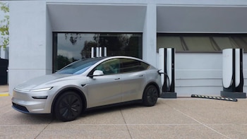 Un automóvil eléctrico plateado está conectado a una estación de carga de color blanco y negro junto a un edificio