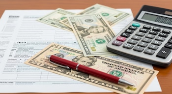 Primer plano de formularios de impuestos, una calculadora gris con pantalla digital y billetes de veinte dólares sobre un escritorio de madera.