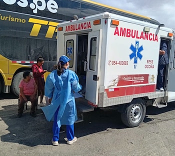 Ambulancia blanca con puertas abiertas, señalización "Ambulancia", persona con equipo de protección, dos mujeres sentadas, autobús y camino de tierra