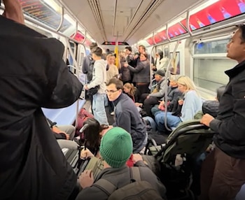 Pasajeros sentados, de pie y agachados en el pasillo interior de un vagón de tren subterráneo con pantallas publicitarias en el techo
