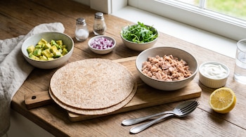 Tortillas de trigo, atún, palta en cubos, cebolla morada, rúcula, yogur griego, medio limón, salero, pimentero, vaso con agua y cubiertos en una mesa de madera.